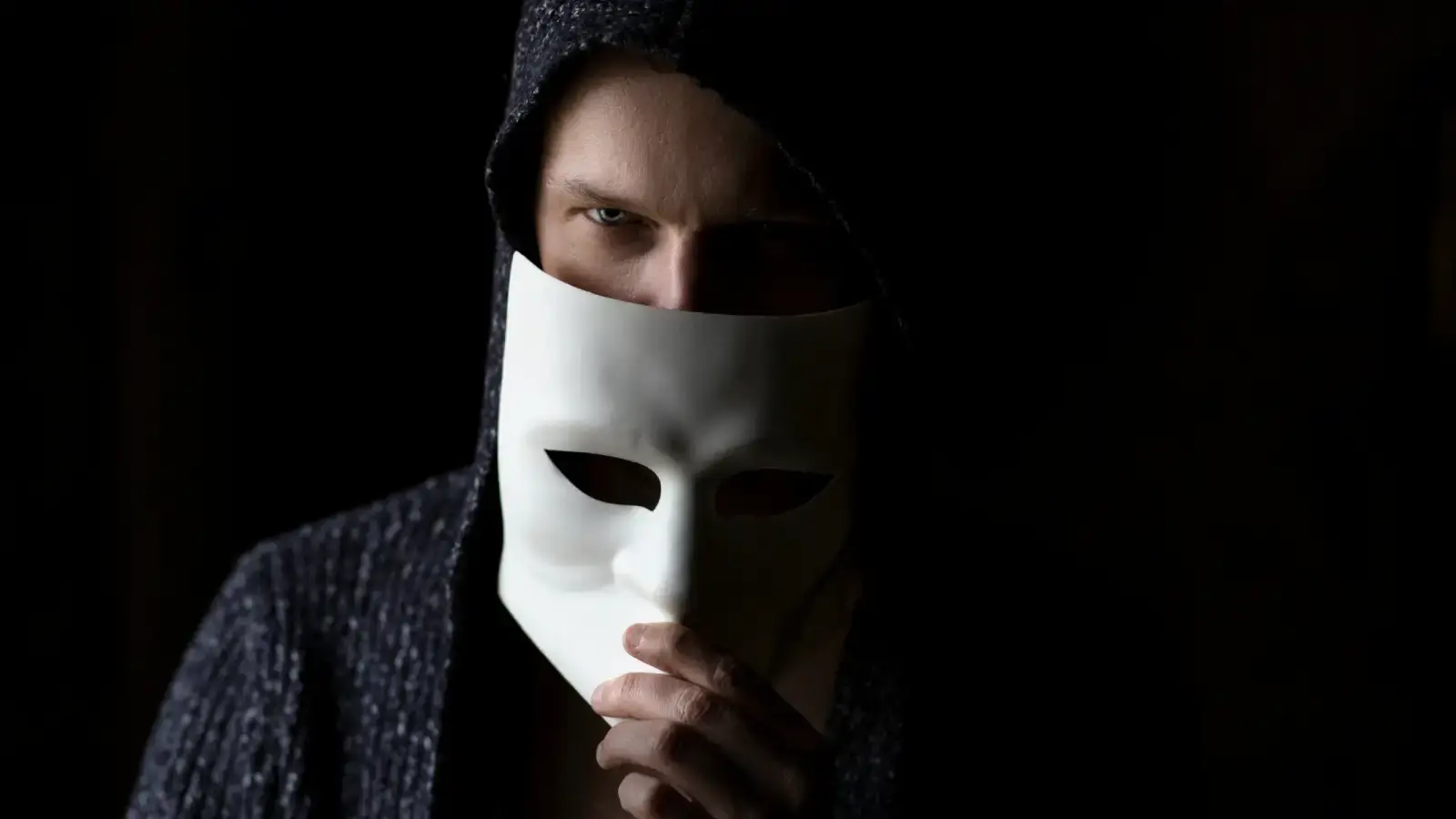 man in dark hoodie holding white mask in front of his face